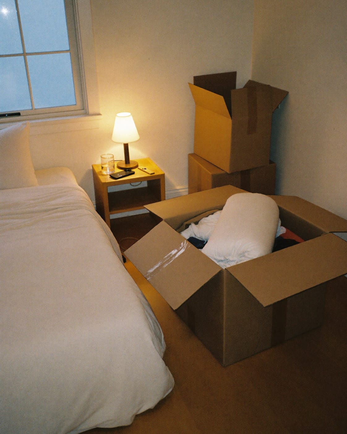 A made bed and a lit bedside lamp with a glass of water and a phone, beside moving boxes, one open with towels and bedding